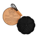 A black silicone waffle mold divided into four heart-shaped sections, paired with a wooden-handled whisk and a round kraft card featuring a waffle recipe.