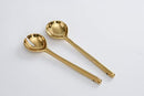 A set of two Pampa Bay salad servers with a lustrous gold titanium-plated finish. One server is a solid spoon, and the other is a slotted spoon or salad fork with two tines. Both have long, slender handles and rest on a light gray surface.