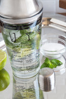 Outset glass cocktail shaker with a stainless steel lid. The glass base is printed with six drink recipes and measurement markings in both ounces and milliliters.