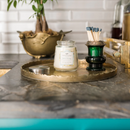 A small, clear glass jar candle with a light beige wax. It has a silver metal lid and a decorative label with the scent "Bella Fresia."