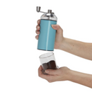 A manual coffee grinder with a clear top and bottom, featuring a vibrant turquoise blue body and a silver hand crank. The background is transparent.