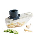 A compact, white and dark gray manual food slicer (mandolin) with a clear container base, shown slicing garlic cloves and cucumber slices.