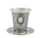 A silver-plated metal Kiddush cup with a textured crosshatch pattern and an ornate, raised oval design on the front. The cup rests on a matching silver-plated saucer.