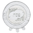 A round, white quilted satin matzah cover with silver embroidery. The design features a decorative frame around the Hebrew word 'Pesach.' The edge has a braided cord trim and three tabs.
