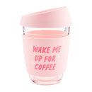 Stylish and practical blush pink travel mug made of glass with a silicone grip and lid, displaying the text "WAKE ME UP FOR COFFEE," from Mud Pie.
