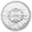 A round, white fabric matzah cover with ornate silver and gold embroidery, a decorative frame, and the Hebrew word 'Pesach' in the center. The edge has a ruffled silver trim.