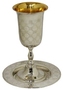 A highly polished, ornate silver kiddush cup with a gold-colored interior, sitting on a matching round silver saucer. Both the cup and the saucer feature a subtle, repeating decorative pattern.
